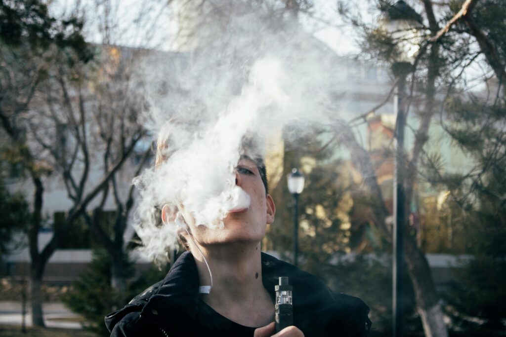 Portrait of a young man vaping in a park, releasing clouds of vapor on a clear day.